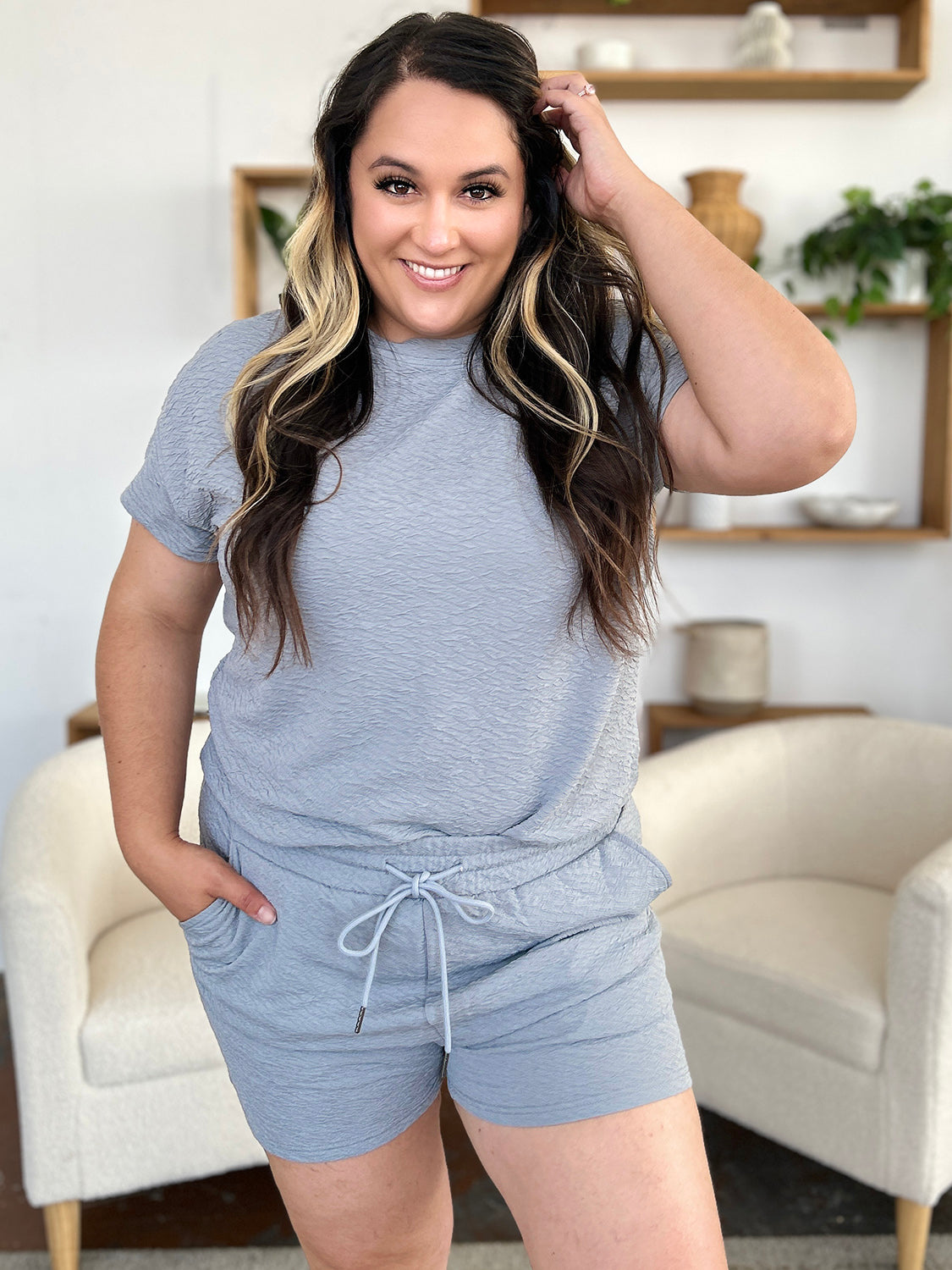 A person with long hair wears the Double Take Full Size Texture Short Sleeve T-Shirt and Drawstring Shorts Set in light gray, accessorized with large earrings and a chain necklace, standing with one hand in their pocket.