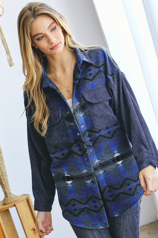 A woman with long blonde hair wears a blue and gray patterned shirt over a white crop top and dark jeans, standing indoors near a window, her casual **Printed Button Down Long Sleeve Jacket** draped casually over her shoulders.