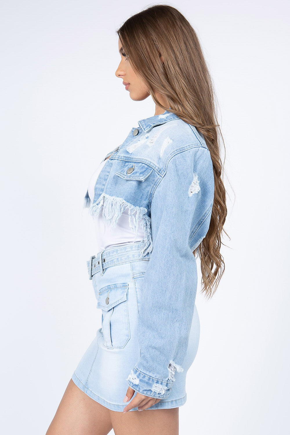 A woman with long brown hair wears an American Bazi Distressed Denim Jacket with Frayed Hem and a light denim skirt with front pockets. The jacket features frayed hems, adding a touch of edgy style. She stands facing the camera against a plain white background.