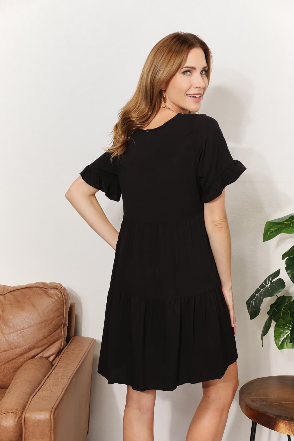 A woman stands indoors wearing the Mandy V-Neck Flounce Sleeve Tiered Dress in navy blue. She has one hand on her hip and is smiling.