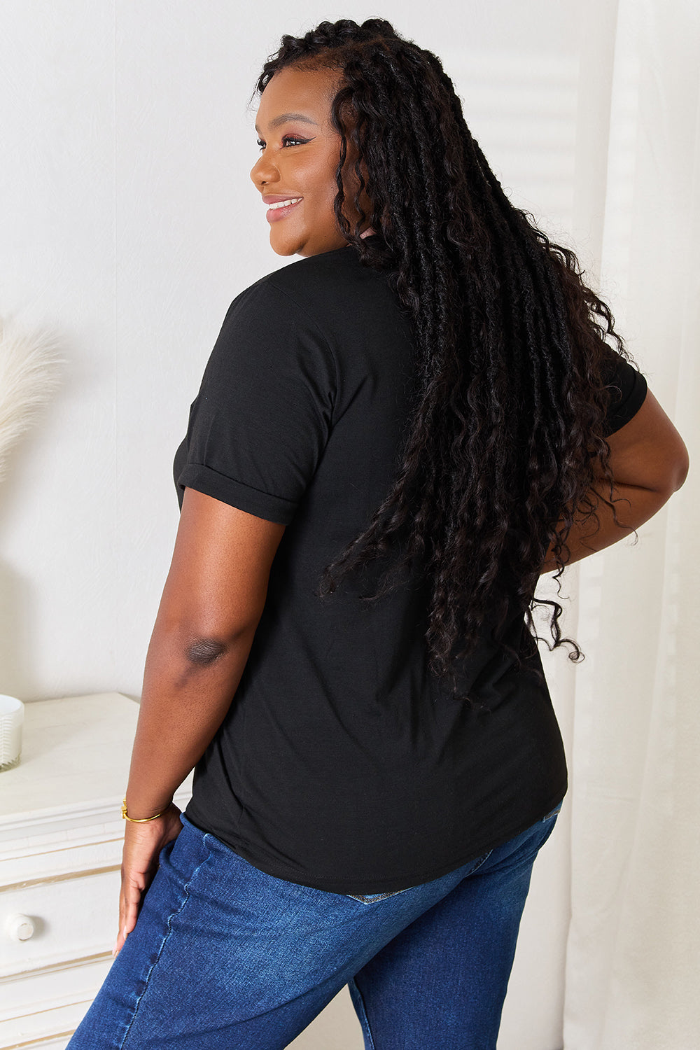 A woman stands indoors wearing the Simply Love Graphic Short Sleeve T-Shirt, a stylish black tee featuring a striking blue and white geometric pattern. Made from comfortable fabric, she has one hand on her hip and is smiling.