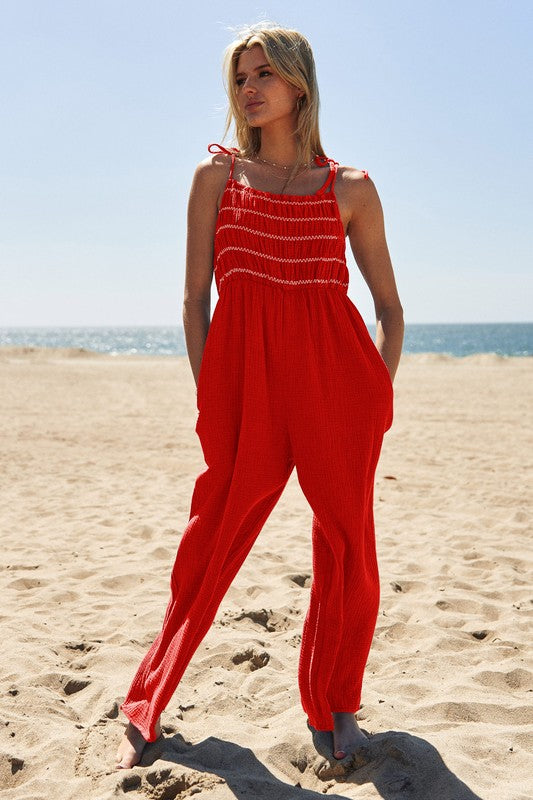 A person wearing the Washed Multi Smocked Detail Tie Straps Jumpsuit stands on the sunny beach, with the ocean creating a perfect backdrop.