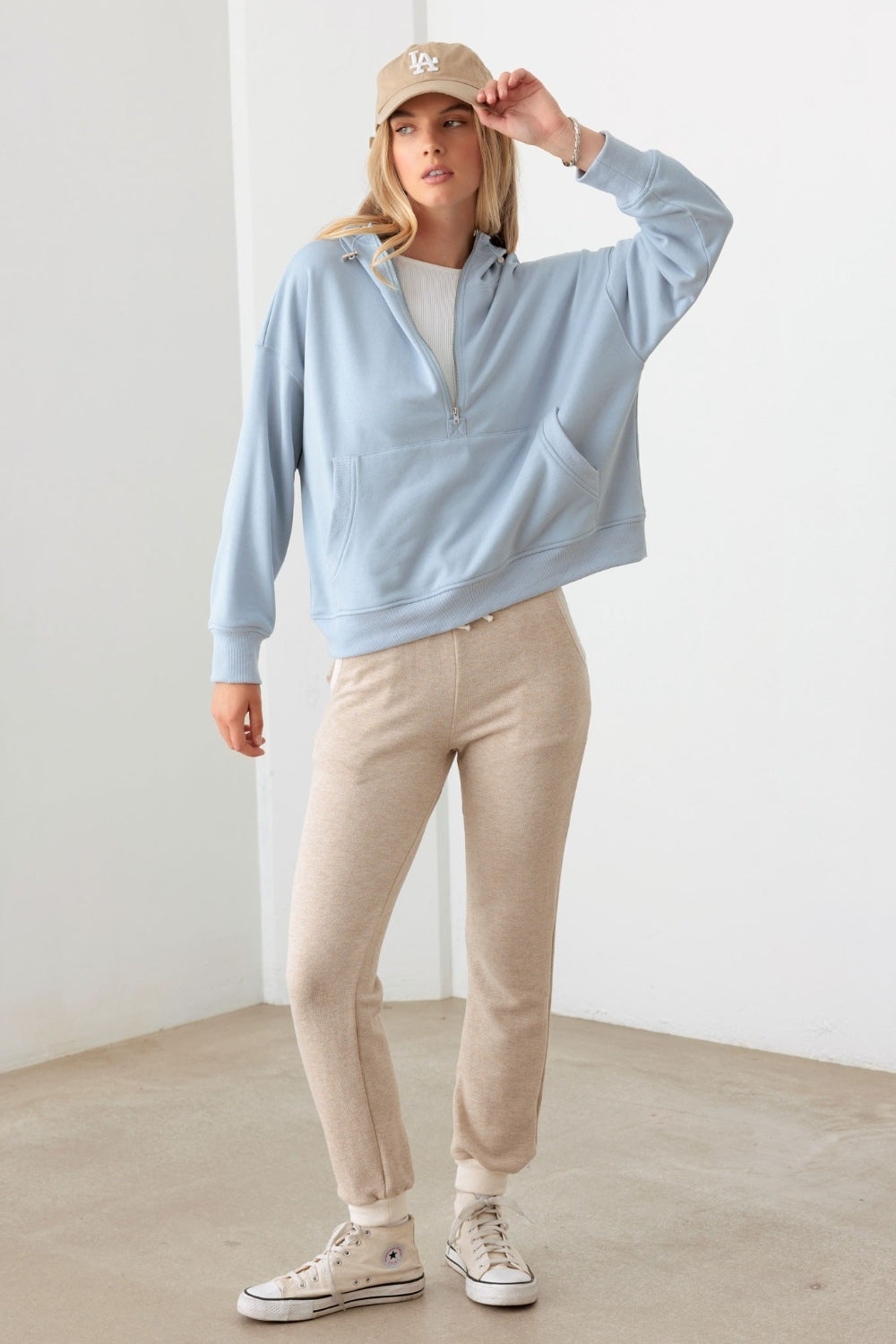 In a neutral-colored room, a person is wearing the Le Lis Half Zip Drawstring Mock Neck Hoodie in light blue along with a beige cap.