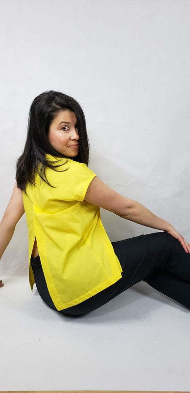 A woman with dark hair is wearing a sleeveless top cotton shirt with front back slits in bright yellow and black pants, seated on the floor against a plain light background, looking over her shoulder.