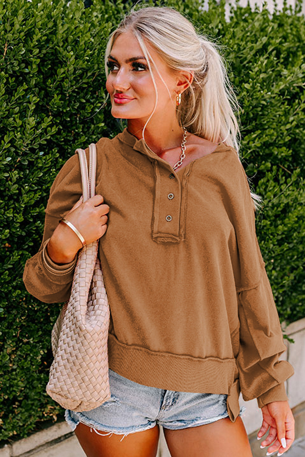 A woman with blonde hair in a ponytail is seen facing away from the camera, standing in front of green bushes while wearing a Chestnut Drop Shoulder Henley Buttons Sweatshirt and denim shorts.