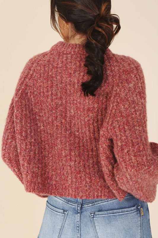 A person is wearing the Melange multicolor sweater top, characterized by textured pink fabric and featuring raglan sleeves along with large buttons, paired with light blue jeans, standing against a plain background.