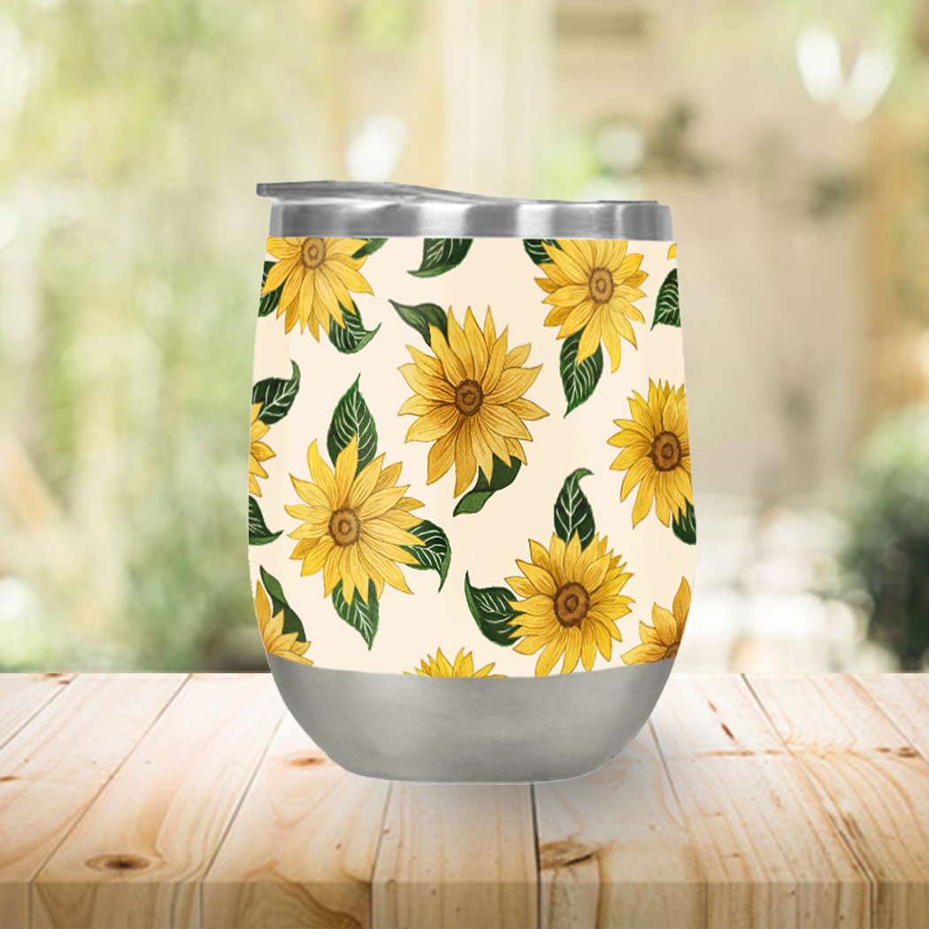 The Summer Sunflower Stemless Wine Tumbler rests on a wooden table against a blurred backdrop, showcasing its stainless steel design with a sunflower pattern and double-wall vacuum insulation. This 12 oz wine tumbler is both BPA and lead-free, guaranteeing each sip is safe as it is stylish.