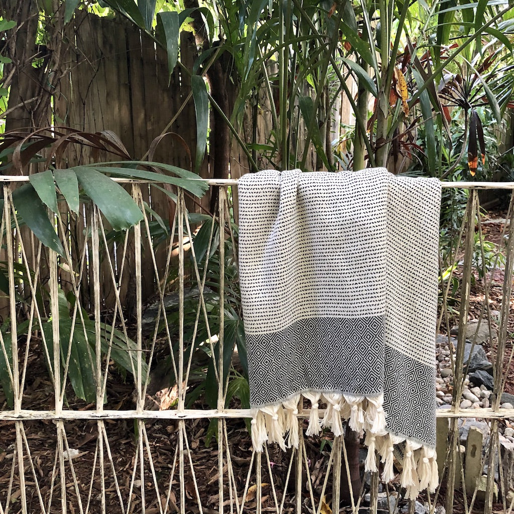 The Diamond Stripe Turkish Towel is crafted from luxurious Turkish cotton and features a folded, patterned design with a black and white striped top section and a red geometric-patterned bottom, complete with tasseled fringe at one end. It's perfect for adding elegance to beach outings or home decor.