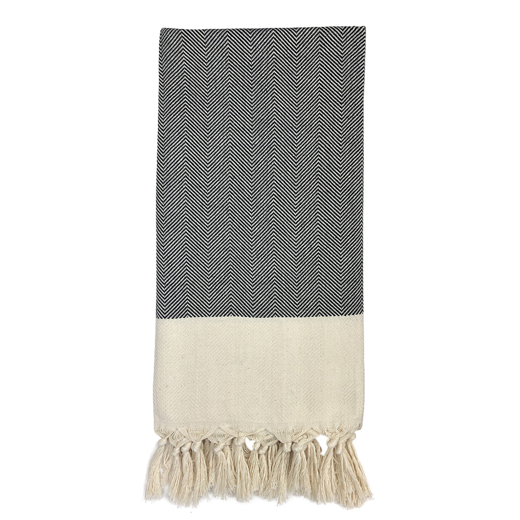 This Herringbone Turkish Towel, crafted from 100% Turkish cotton and adorned with a black and white chevron pattern, features hand-knotted fringes elegantly laid flat against a white background.