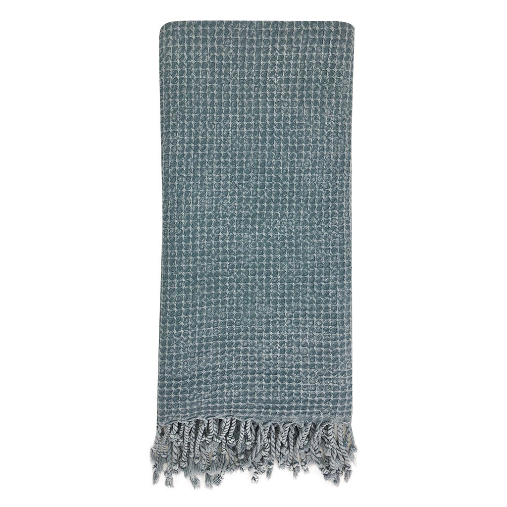 A folded, light gray blanket featuring a waffle texture and fringe tassels at the bottom mirrors the elegance of the Waffle Weave Turkish Towel, combining style and practicality for any cozy setting.