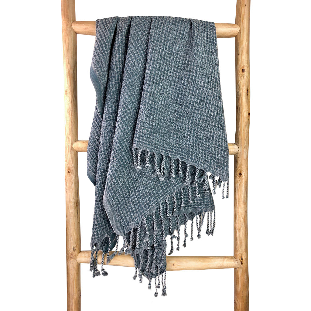 A folded, light gray blanket featuring a waffle texture and fringe tassels at the bottom mirrors the elegance of the Waffle Weave Turkish Towel, combining style and practicality for any cozy setting.