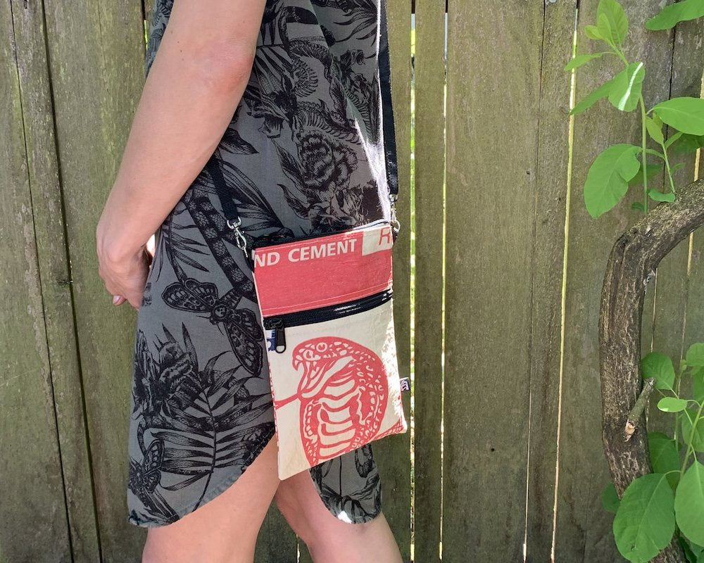 The Breezy Crossbody showcases a vibrant blue fish print on a pink background, crafted from upcycled materials. It includes two black zippered compartments and an adjustable black strap.