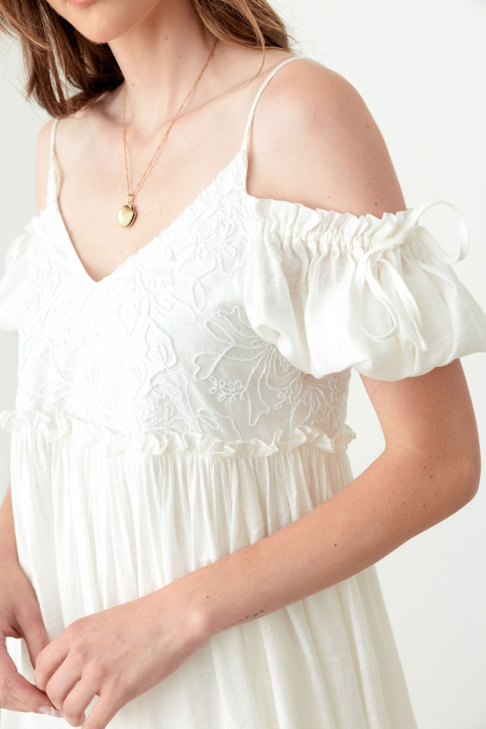 A woman poses in a boho-inspired, embroidered floral shirred cold shoulder mini dress by Mustard Seed. She accessorizes with a gold necklace and maintains a neutral expression against the backdrop of a plain white wall.