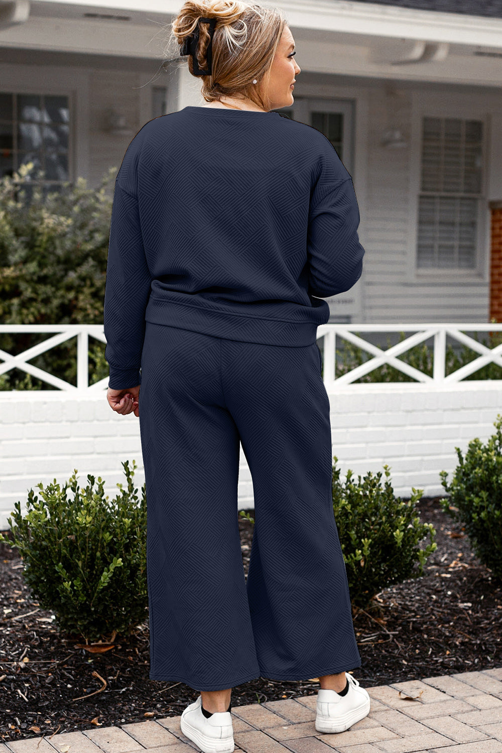 A woman with blonde hair, dressed in the Double Take Full Size Textured Long Sleeve Top and Drawstring Pants Set featuring a stretchy black long-sleeve top and matching wide-leg trousers made of rayon spandex, stands barefoot in a room with white walls and a wooden floor.