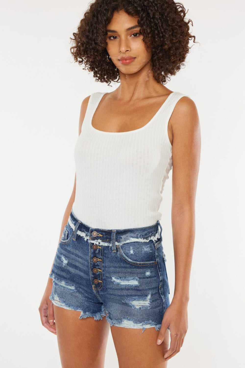 A woman with curly hair wearing a white sleeveless top and Kancan Distressed Button Fly Denim Shorts stands against a plain background, perfect for a stylish summer wardrobe.