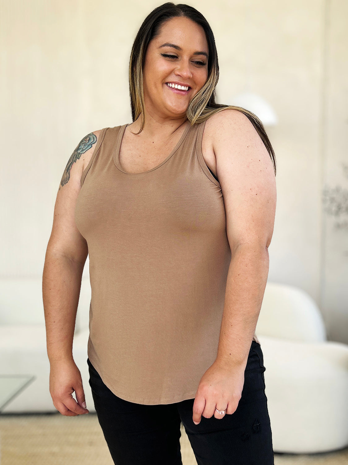 A woman wearing sunglasses, blue jeans, and the Basic Bae Full Size Round Neck Curved Hem Tank is standing in front of a concrete wall, holding her glasses with one hand and carrying a beige shoulder bag. Her basic style complements the slightly stretchy fabric of her outfit, ensuring comfort while maintaining chic simplicity.