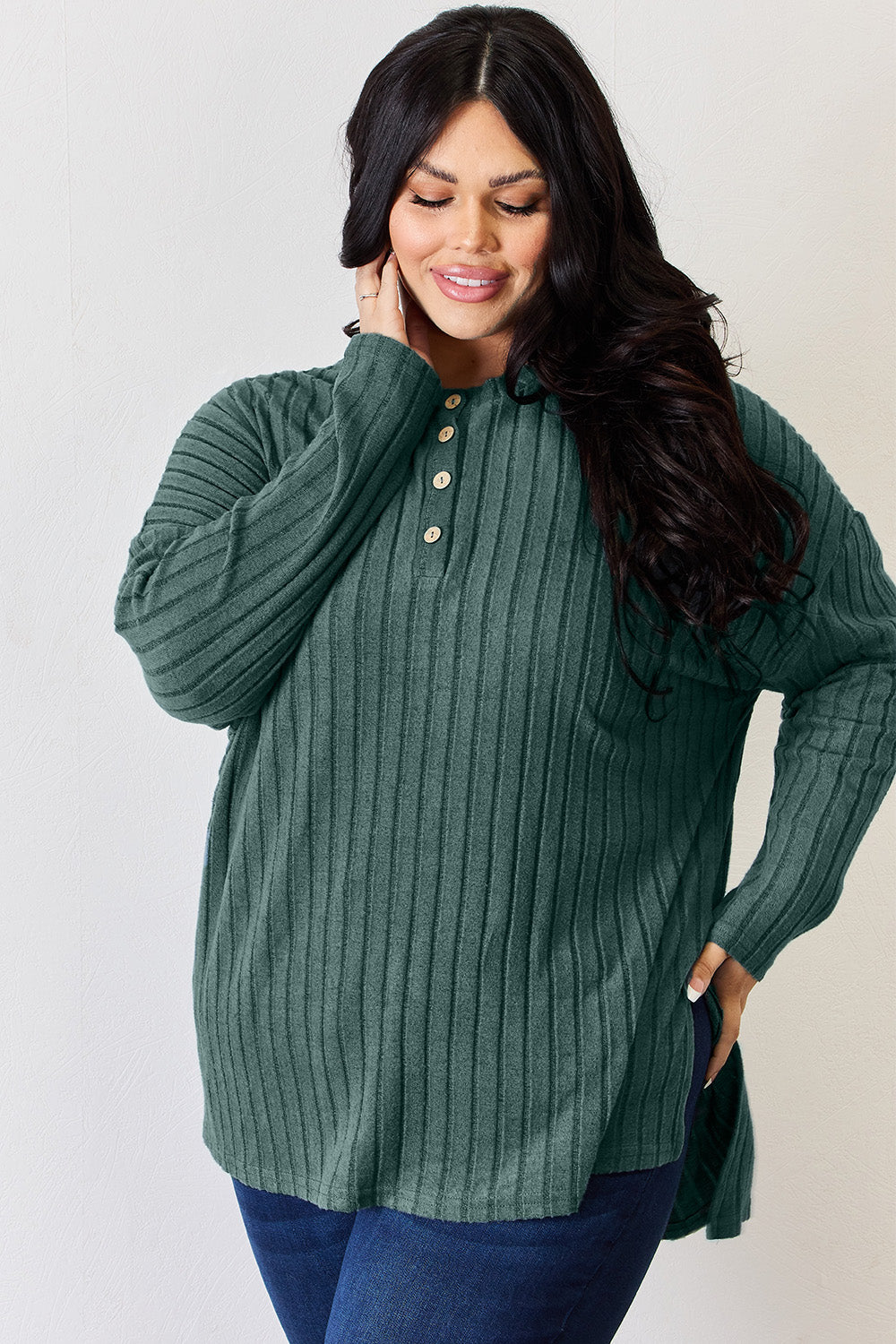 A person with long blonde hair is standing against a light background, wearing the Basic Bae Full Size Ribbed Half Button Long Sleeve High-Low T-Shirt in blue and dark pants. They are looking at the camera and smiling slightly, showcasing the chic high-low hem of their top.