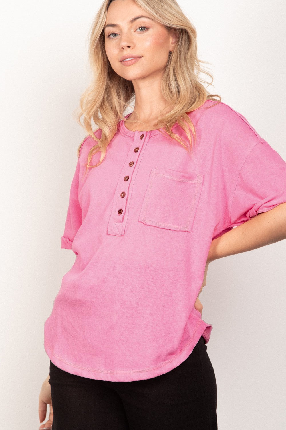 A person with long blonde hair is wearing the VERY J Twisted Sleeve Band Half Button Top in pink, standing in front of a plain white background.