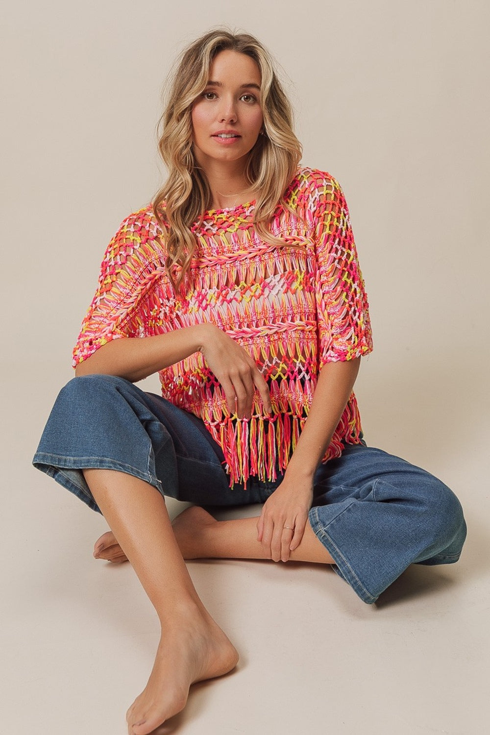 A person with long hair wears the BiBi Openwork Fringed Knit Cover Up, exuding bohemian flair, paired with jeans. They sit with one hand raised and the other resting on their lap against a plain background.