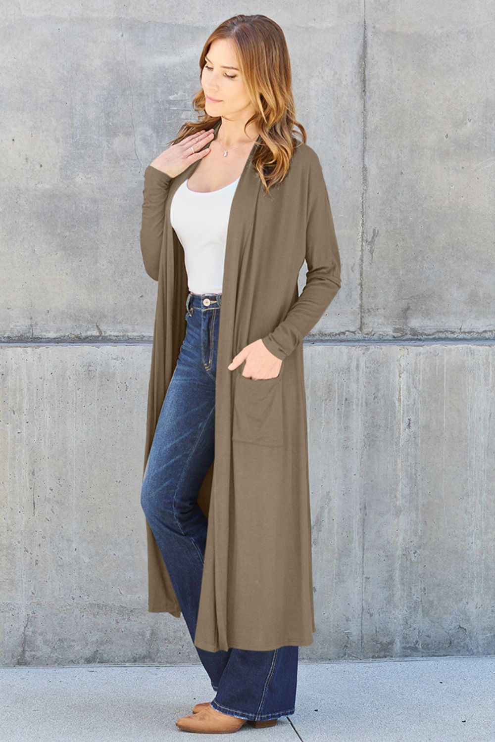 A woman stands against a concrete wall, wearing the Basic Bae Full Size Open Front Long Sleeve Cover Up made from rayon spandex, a white top, blue jeans in her perfect size, and brown shoes with her hands in her pockets. Machine wash cold for best results.