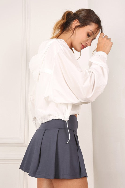 A woman wearing a pink wind breaker over a white top stands against a white wall.