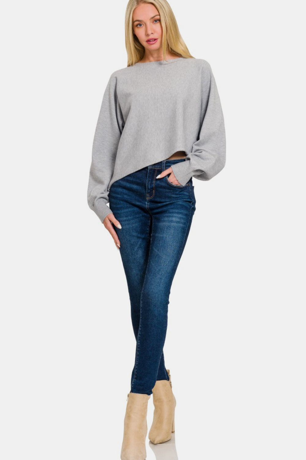 A person with long blonde hair wears a stylish Zenana Asymmetric Hem Long Sleeve Sweater made of soft and comfortable gray fabric, paired with blue jeans, standing against a plain white background.