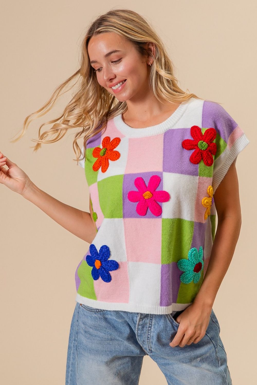 A woman sits in a rattan hanging chair, wearing a BiBi Flower Patch Checkered Sweater Vest with denim shorts. She has long blonde hair and is looking to the side, with her hand resting on her head, epitomizing a cozy layered ensemble perfect for a trendy fall wardrobe.