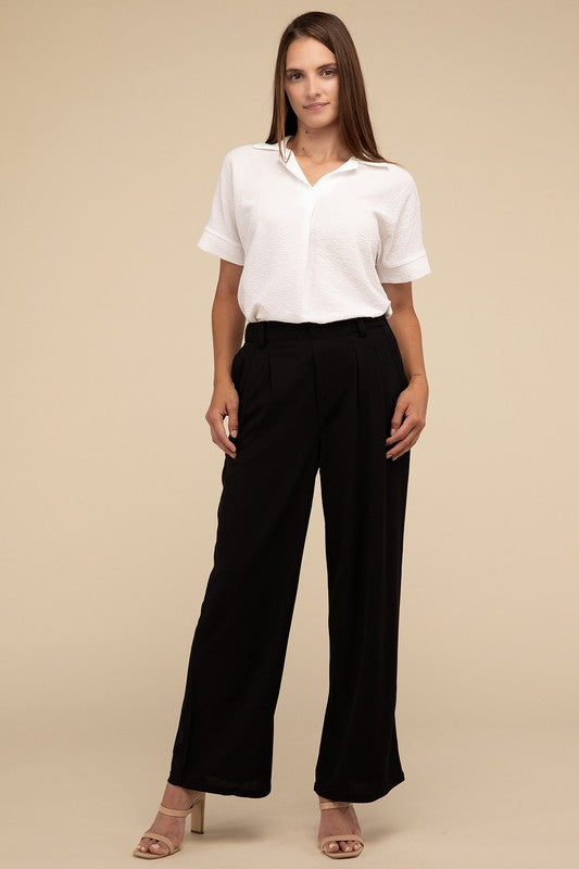 A woman stands against a beige background, highlighting her chic ensemble with a tailored fit. She wears a white short-sleeve shirt paired with versatile pink Waffle Trouser Pants, hands casually in pockets, complemented by stylish strappy sandals.