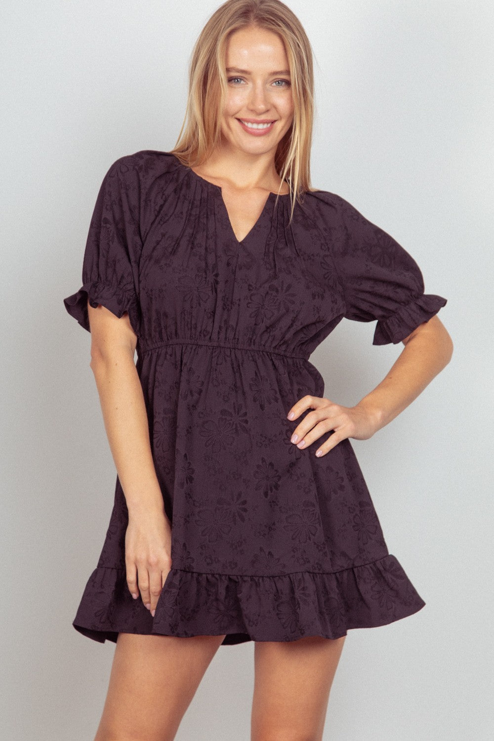 Person in a VERY J Floral Textured Woven Ruffled Mini Dress standing and smiling with one hand on hip.