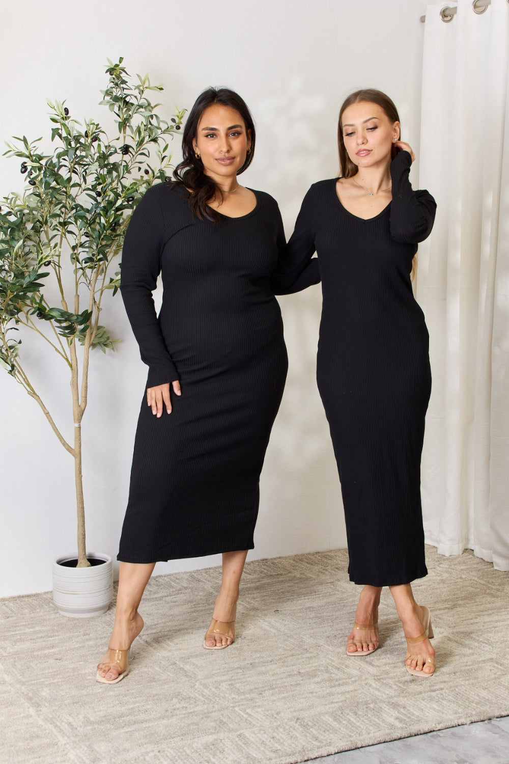 A person stands indoors wearing the chic Culture Code Full Size Ribbed Long Sleeve Midi Slit Dress paired with sandals, next to a potted plant.