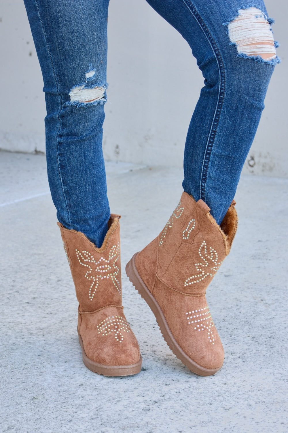 A person wearing ripped blue jeans and Forever Link Rhinestone Furry Flat Boots stands on a concrete surface.