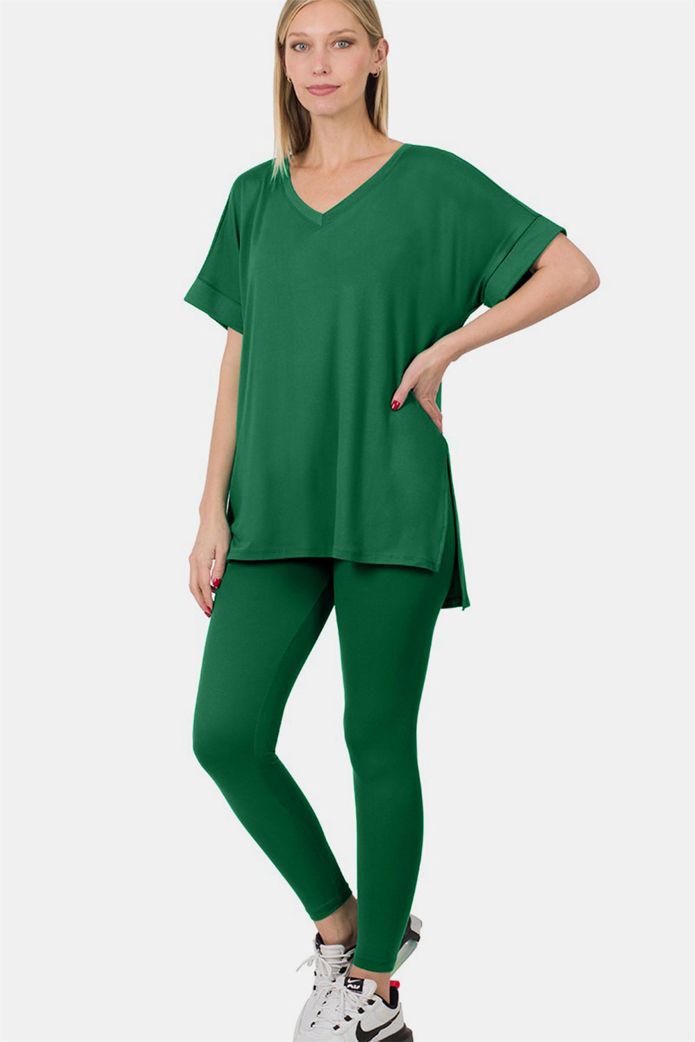 A person wearing the Zenana V-Neck Rolled Short Sleeve T-Shirt and Leggings Lounge Set in green, standing and smiling with one hand touching the side of their head, stylishly matched with sandals.