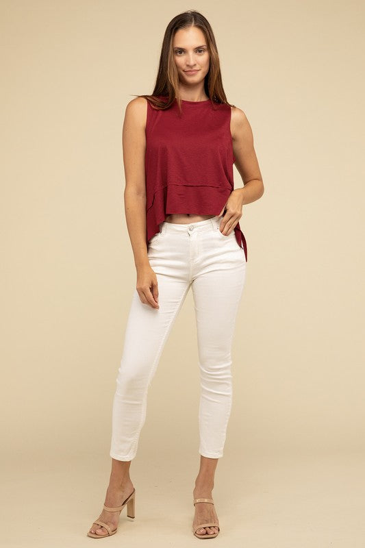 Wearing the Shark Bite Side Slit Short Sleeveless Top in maroon paired with white pants, a model is set against a beige background, exemplifying an effortlessly chic and casual wardrobe.