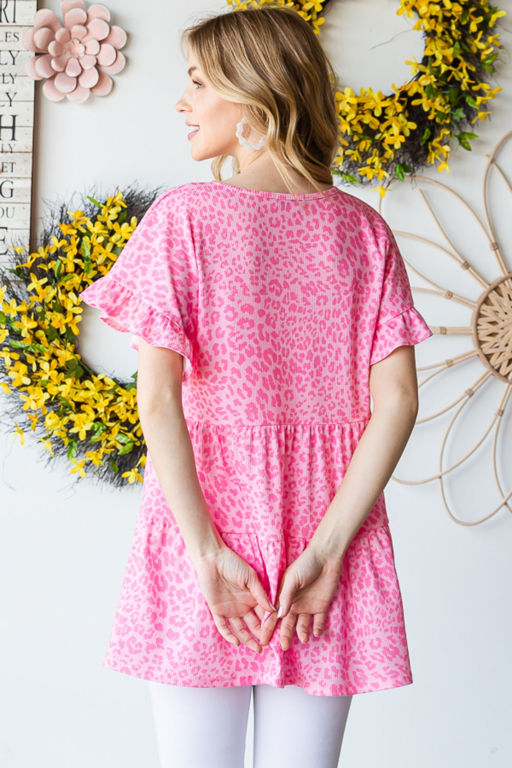 A person wearing a Heimish Full Size Leopard Ruffle Trim Short Sleeve Top stands in front of a floral wreath display.