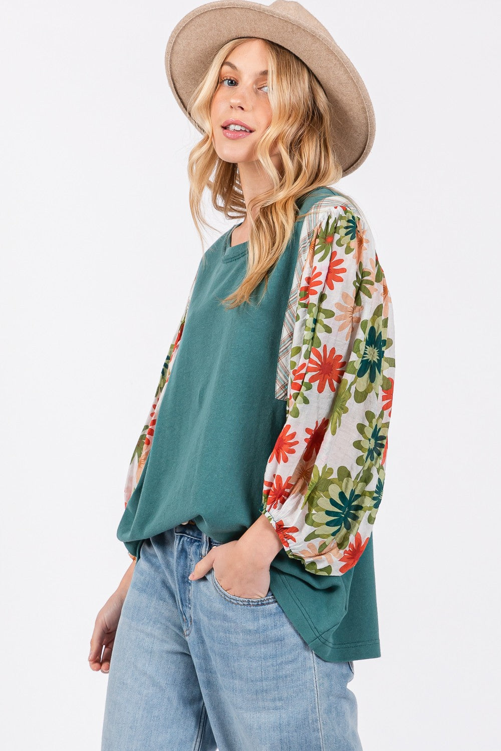 A person in a hat wears the SAGE + FIG Full Size Printed Balloon Sleeve Contrast Top with teal floral-patterned sleeves, blue jeans, and a back heart patch detail.