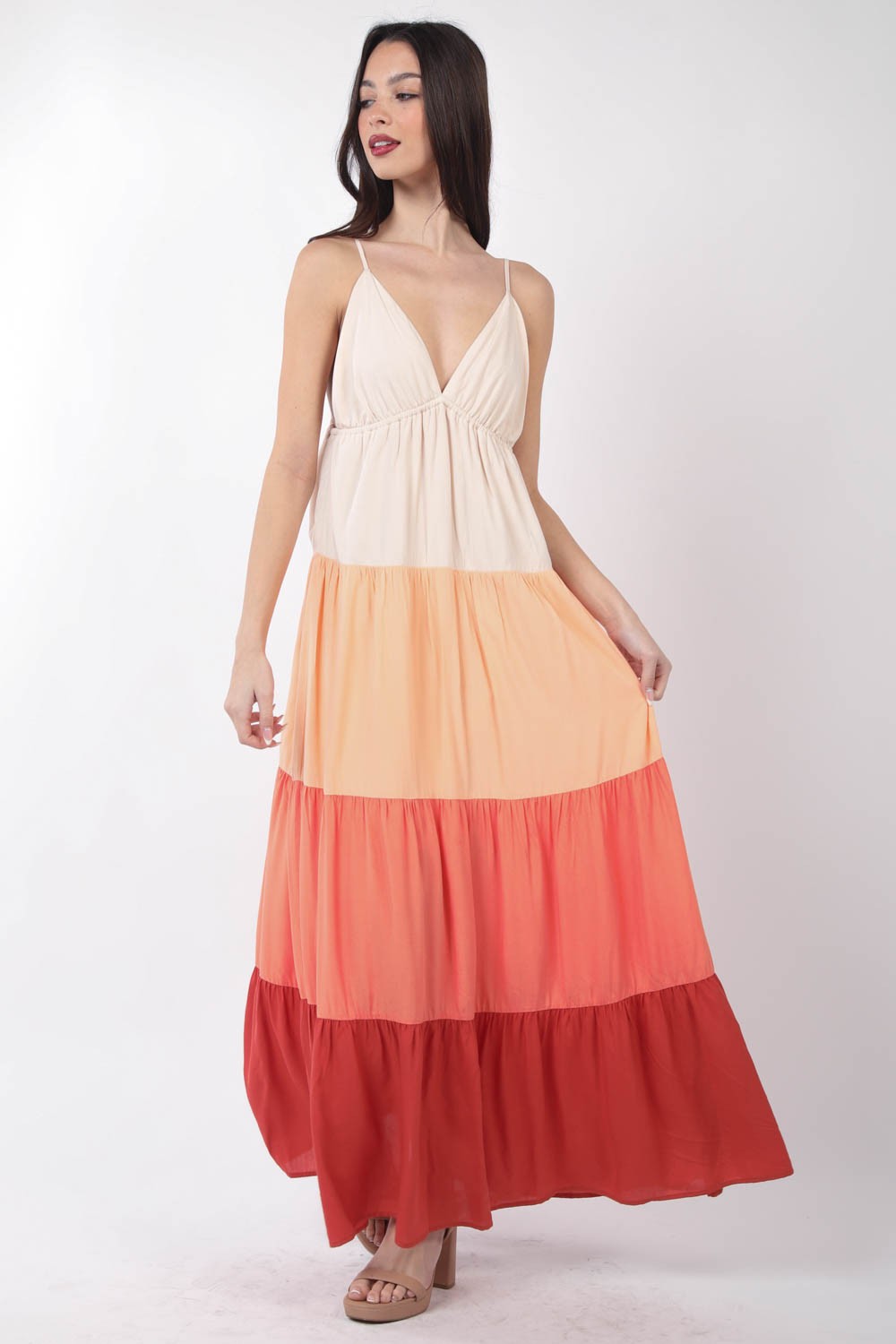 A woman stands, wearing the VERY J Color Block Tiered Maxi Cami Dress in shades of beige, peach, and red, paired with beige sandals. Her right hand holds the side of her dress as she poses against a plain white background—a truly fashionable choice.