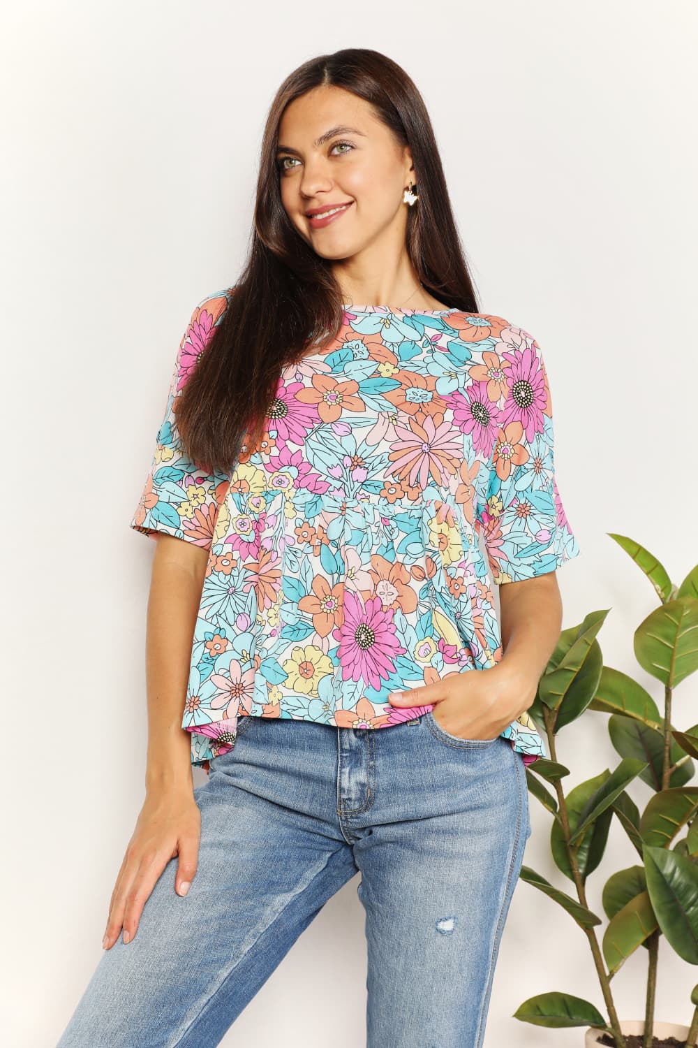 A person wearing a Double Take Floral Round Neck Babydoll Top and jeans stands with one hand in their pocket, with a plant in the background.