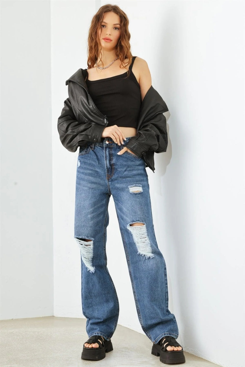 Person wearing HAMMER COLLECTION Distressed High Waist Jeans with ripped knees and black platform sandals, standing in a minimalist setting. The flattering fit and edgy distressed details add to the overall chic look.