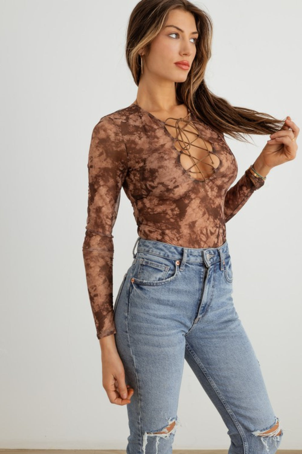 A woman with long hair is wearing the HERA COLLECTION Abstract Mesh Lace-Up Long Sleeve Bodysuit paired with ripped blue jeans, creating a statement-making look as she stands against a plain background.