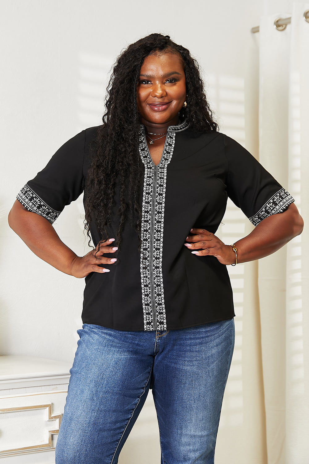 Wearing the elegant Double Take Embroidered Notched Neck Top and blue jeans, a woman with long, wavy hair stands smiling against a light background.