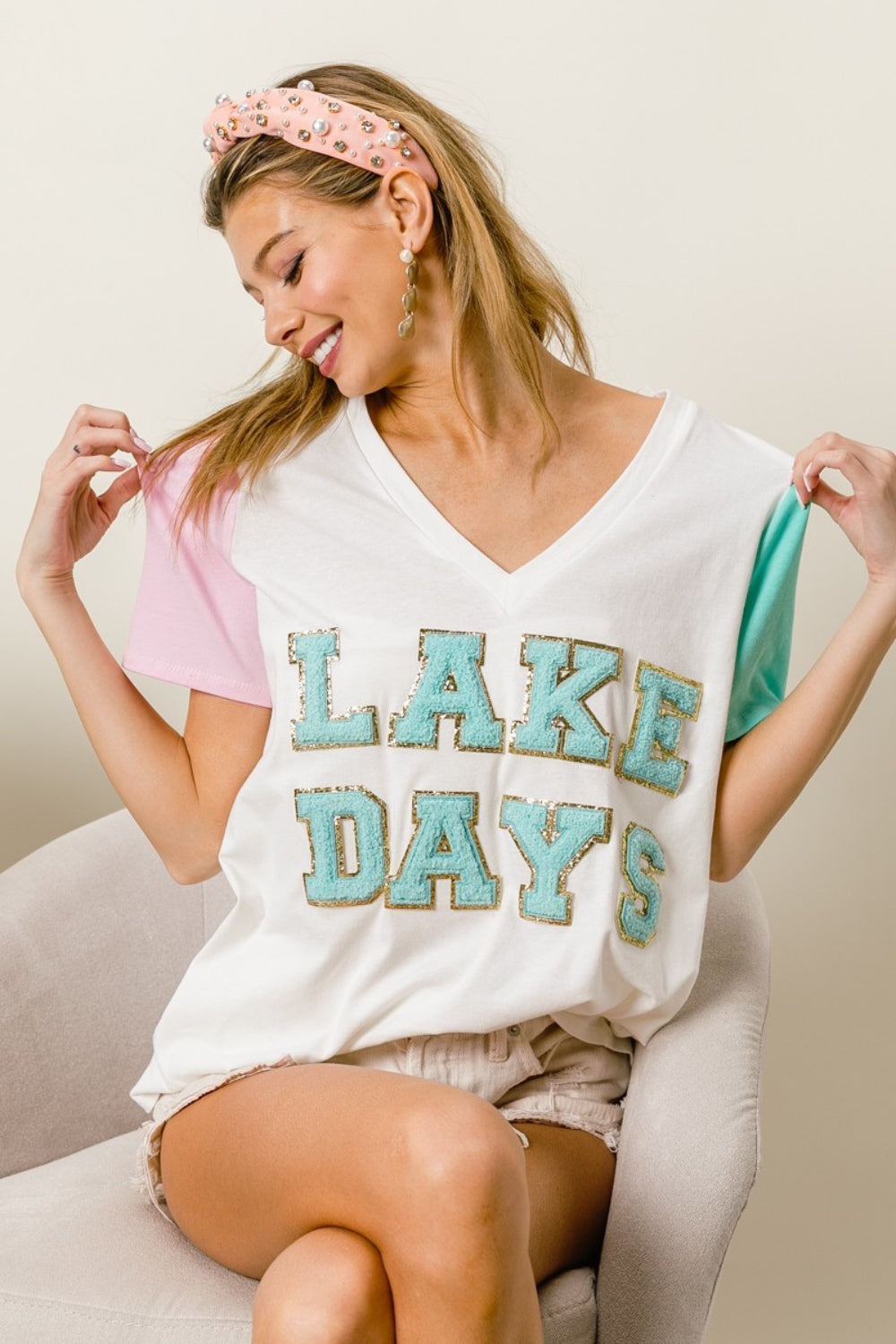 A woman wearing a BiBi Letter Patch V-Neck Color Block Short Sleeve T-Shirt, which features "Lake Days" printed on it, sits on a chair. She holds the shirt's sleeves and wears a headband and earrings.