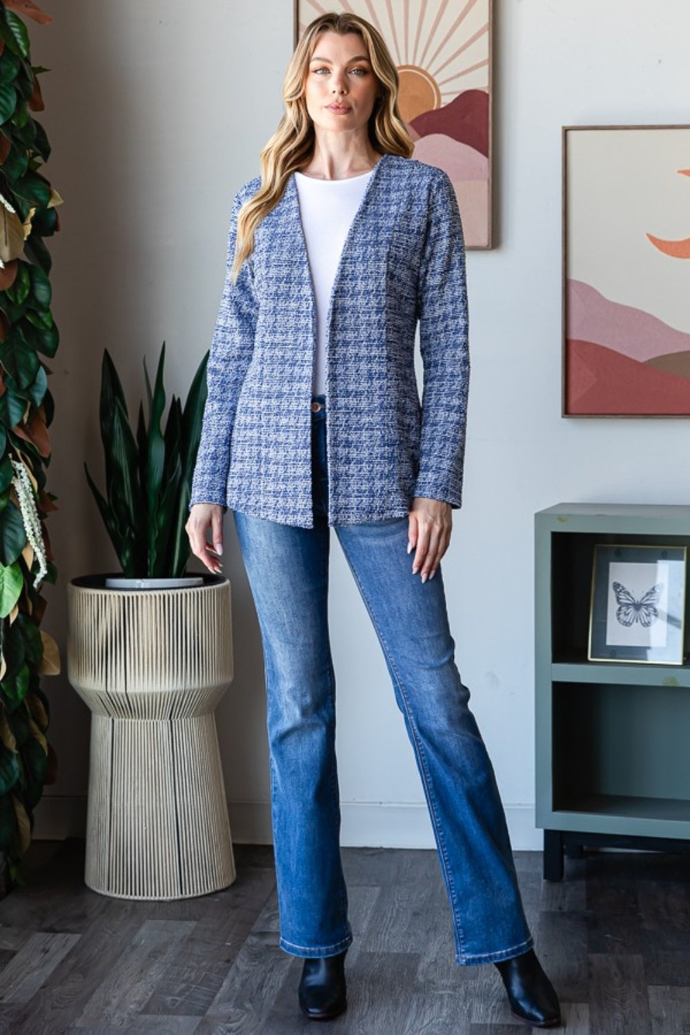 A woman with wavy blonde hair wears the Heimish Full Size Houndstooth Tweed Open Front Blazer in blue, paired with a white top and blue jeans. She stands in front of a wall adorned with framed art.