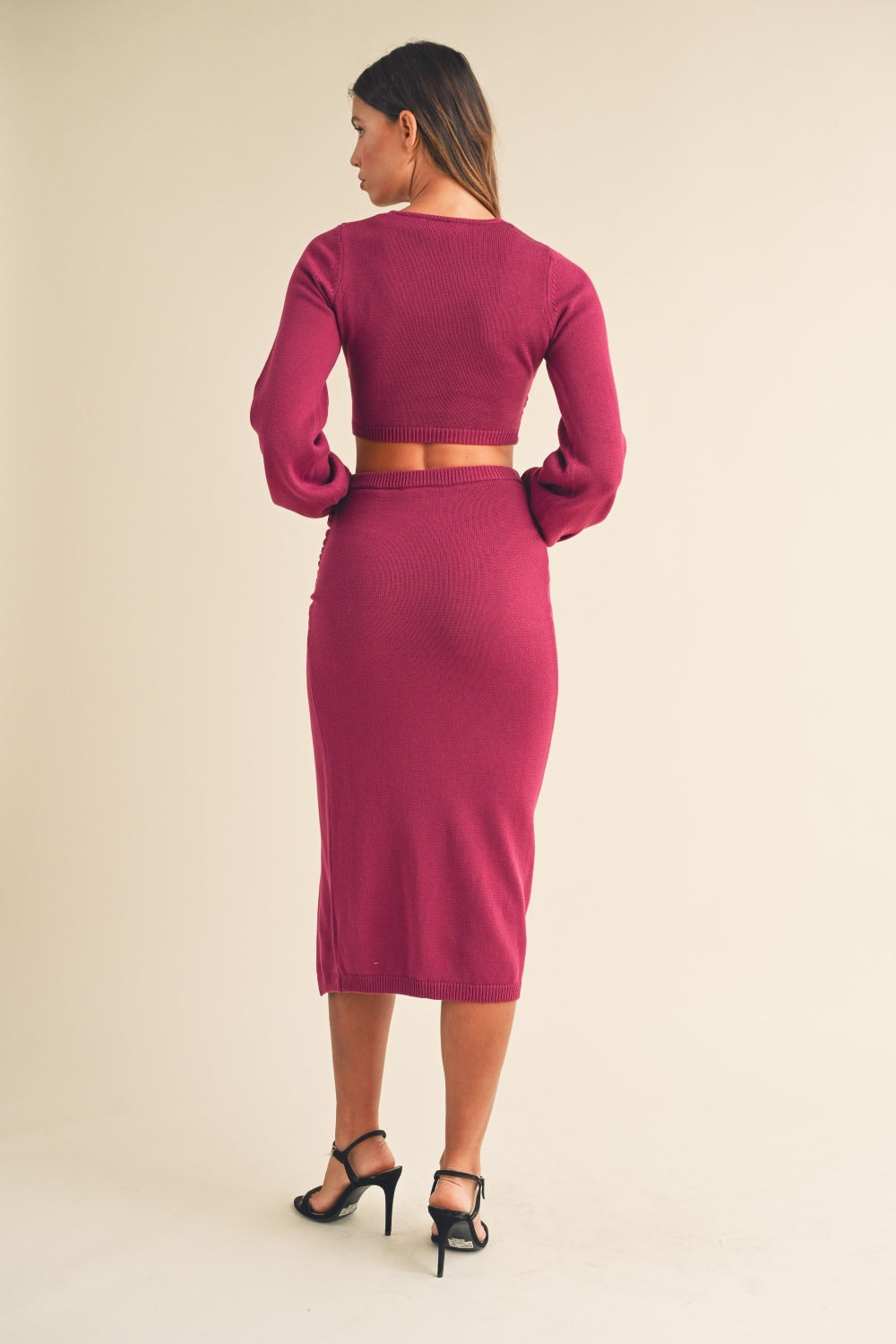 A woman wearing the MABLE Front Twisted Knit Top and Midi Skirt Set, featuring a long-sleeved, magenta knitted top with a front twist detail and a matching high-waisted, thigh-slit midi skirt, stands against a plain background.