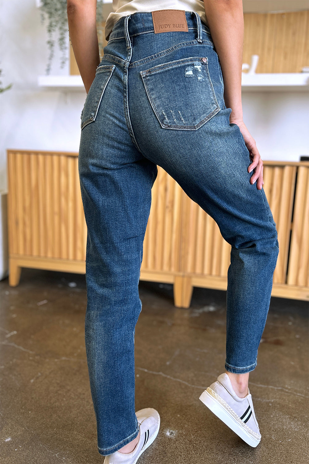 Person wearing Judy Blue Full Size Tummy Control High Waist Slim Jeans in blue and a white shirt, with their hands in their pockets. They are standing indoors on a polished concrete floor with a wooden cabinet and plant in the background.