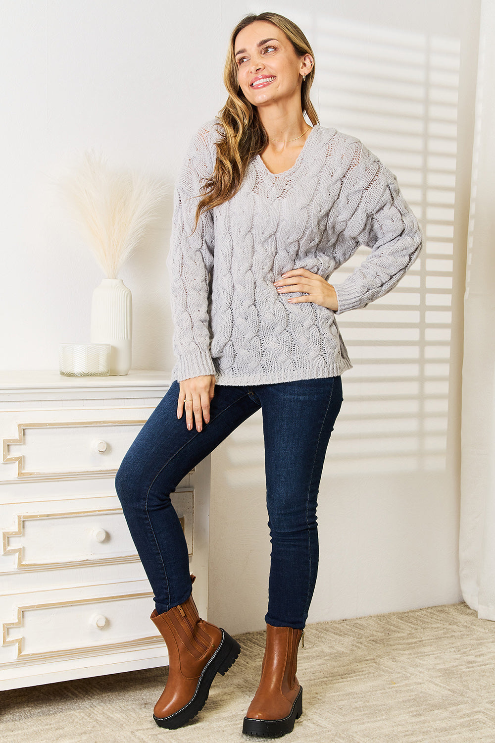 A person with long hair wearing a cozy Woven Right Cable-Knit Hooded Sweater and dark jeans is standing indoors looking to the side.