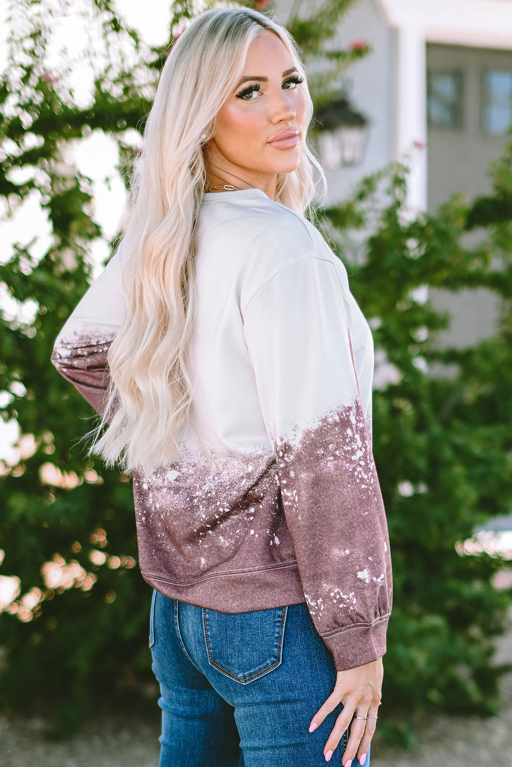 A person with long blonde hair is standing outdoors, facing away from the camera, wearing a Tie Dye Gradient Pullover Sweatshirt featuring a white and brown ombre splatter design.