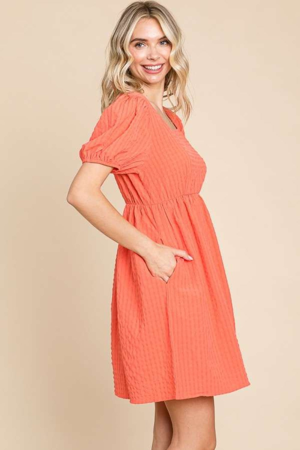 A woman is wearing a Culture Code Textured Square Neck Short Sleeve Dress: it's a stylish, short-sleeved, square-necked, orange dress with puffed sleeves. She is smiling and has one hand in her pocket.