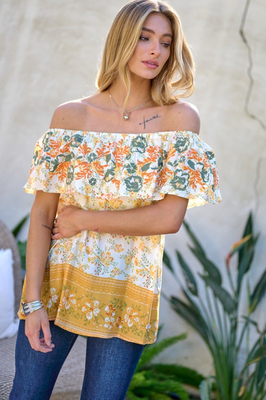 A woman with blonde hair is smiling while standing outdoors, wearing the Printed Off Shoulder Smocked Top paired with jeans.