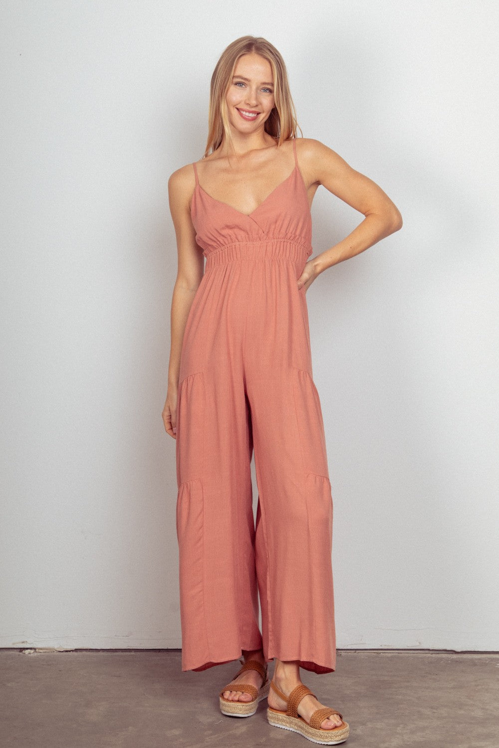 A woman stands against a plain background, wearing the VERY J Sleeveless Ruched Wide Leg Jumpsuit in pink, featuring an elastic waist and adjustable straps. She completes the look with crossover sandals.