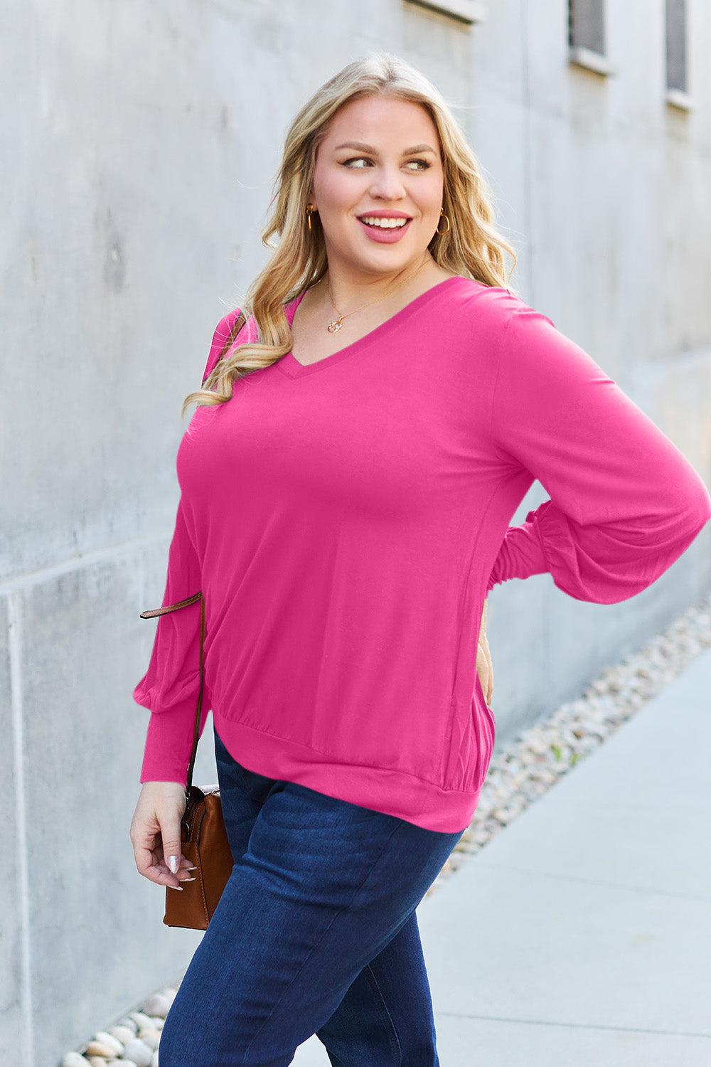 A woman with brown hair is standing against a concrete wall, wearing the Basic Bae Full Size V-Neck Lantern Sleeve Top in teal and blue basic-style jeans, smiling.